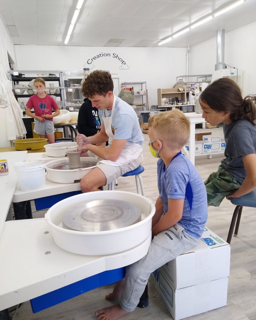 Young man showing little kids how to make clay artwork on the pottery wheel.